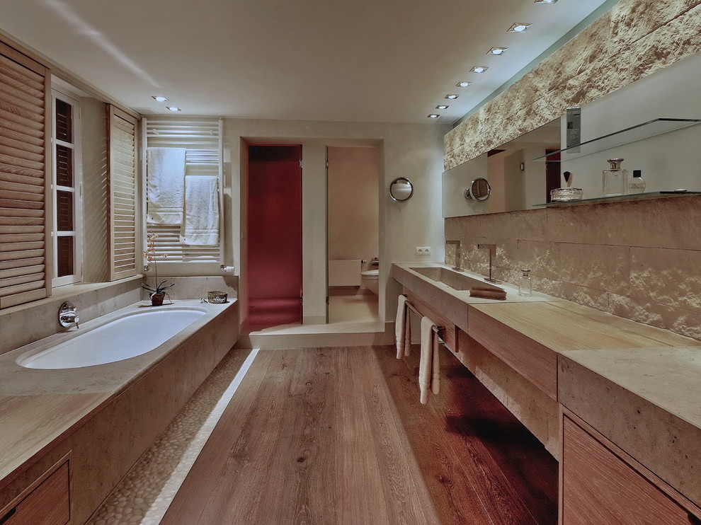 Immagine di una stanza da bagno minimal di medie dimensioni con piastrelle in pietra, pavimento in legno massello medio, lavabo integrato, top in cemento, vasca sottopiano e porta doccia a battente