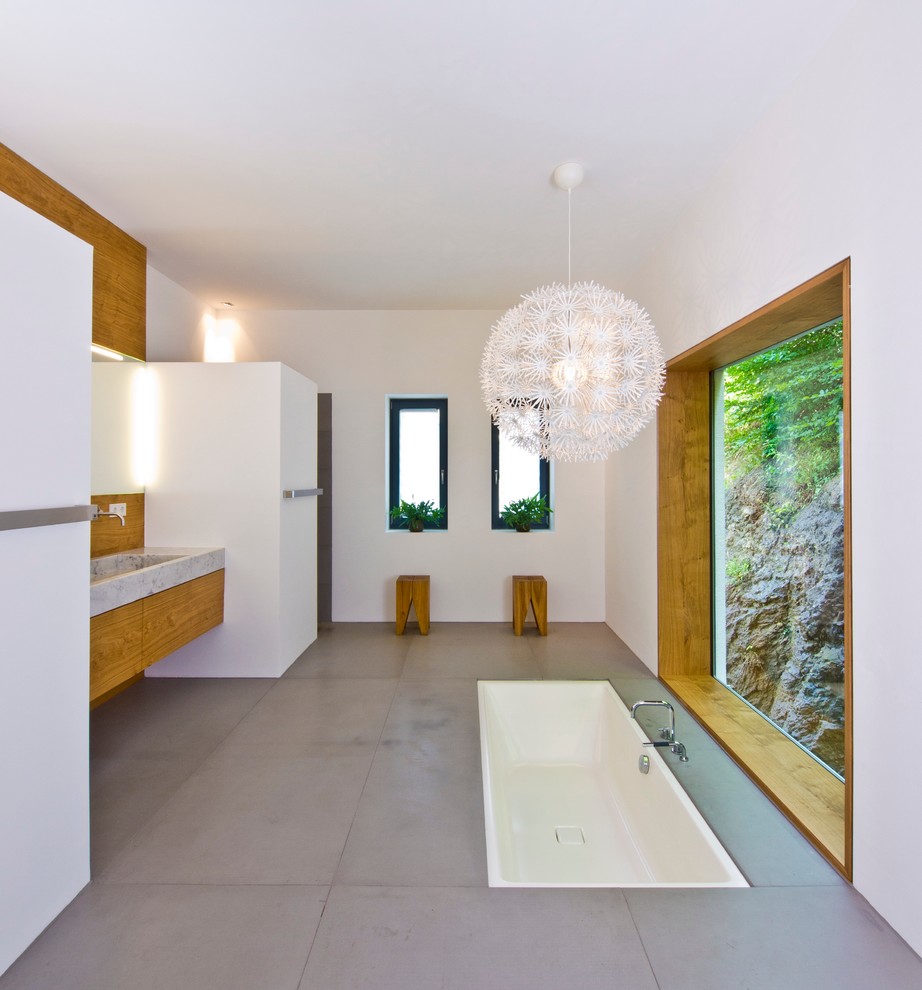 На фото: большая ванная комната в стиле модернизм с полновстраиваемой ванной, плоскими фасадами, фасадами цвета дерева среднего тона, белыми стенами, бетонным полом, монолитной раковиной, мраморной столешницей и окном