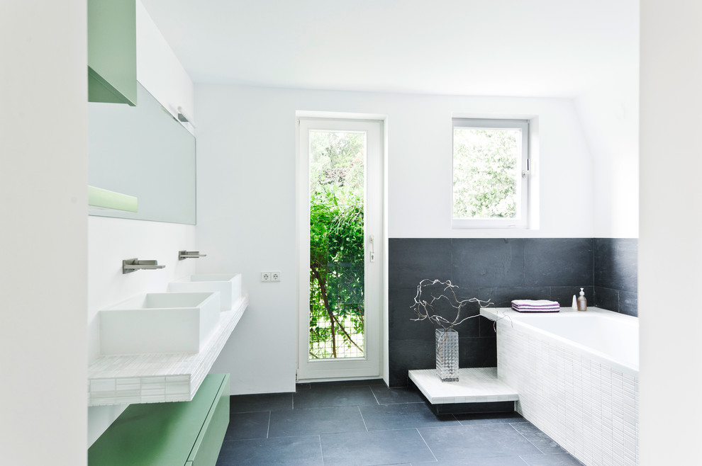 Bild på ett mellanstort skandinaviskt badrum, med ett fristående handfat, släta luckor, gröna skåp, kaklad bänkskiva, svart kakel, vita väggar, ett platsbyggt badkar, stenhäll och skiffergolv