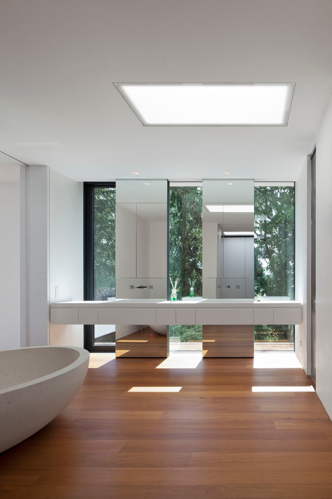 Modern inredning av ett mellanstort badrum, med ett fristående badkar, spegel istället för kakel, släta luckor, vita skåp, vita väggar och mellanmörkt trägolv
