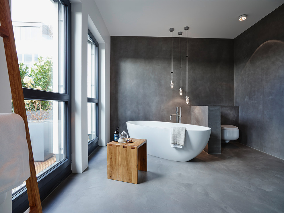 Cette photo montre une grande salle de bain principale tendance avec une baignoire indépendante, WC suspendus, un mur gris, sol en béton ciré et une fenêtre.