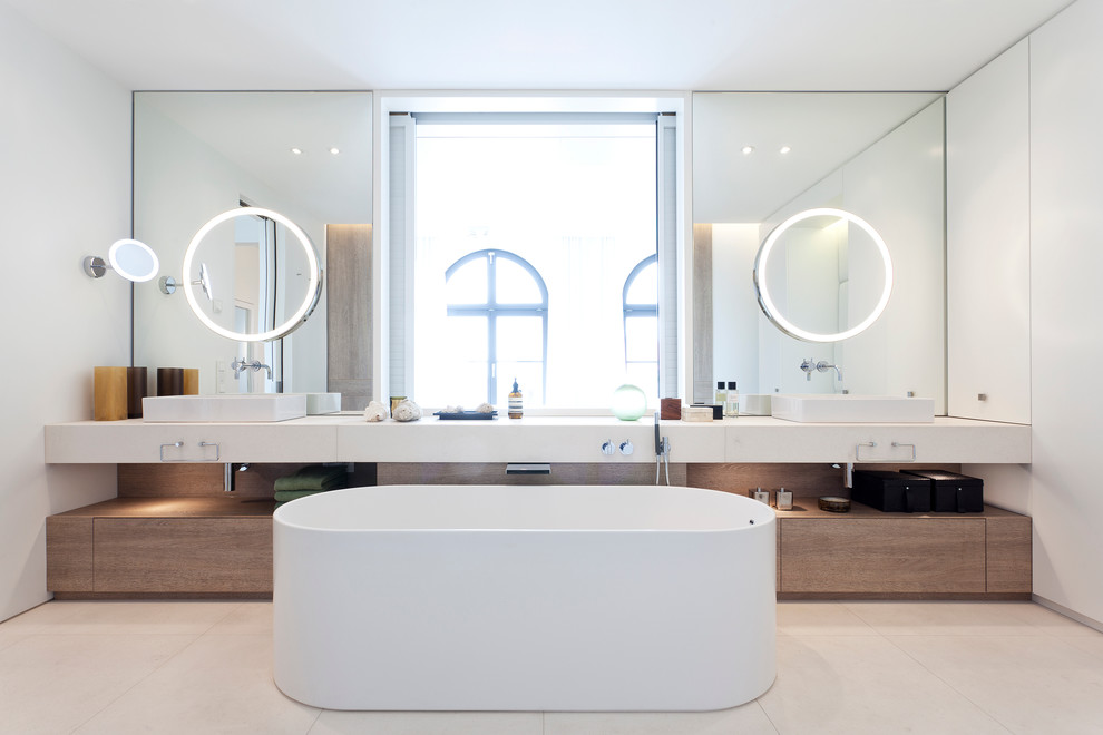 Идея дизайна: огромная главная ванная комната в современном стиле с плоскими фасадами, белыми фасадами, отдельно стоящей ванной, душем без бортиков, инсталляцией, бежевой плиткой, плиткой из листового камня, белыми стенами, полом из известняка, настольной раковиной, столешницей из известняка, окном и зеркалом с подсветкой