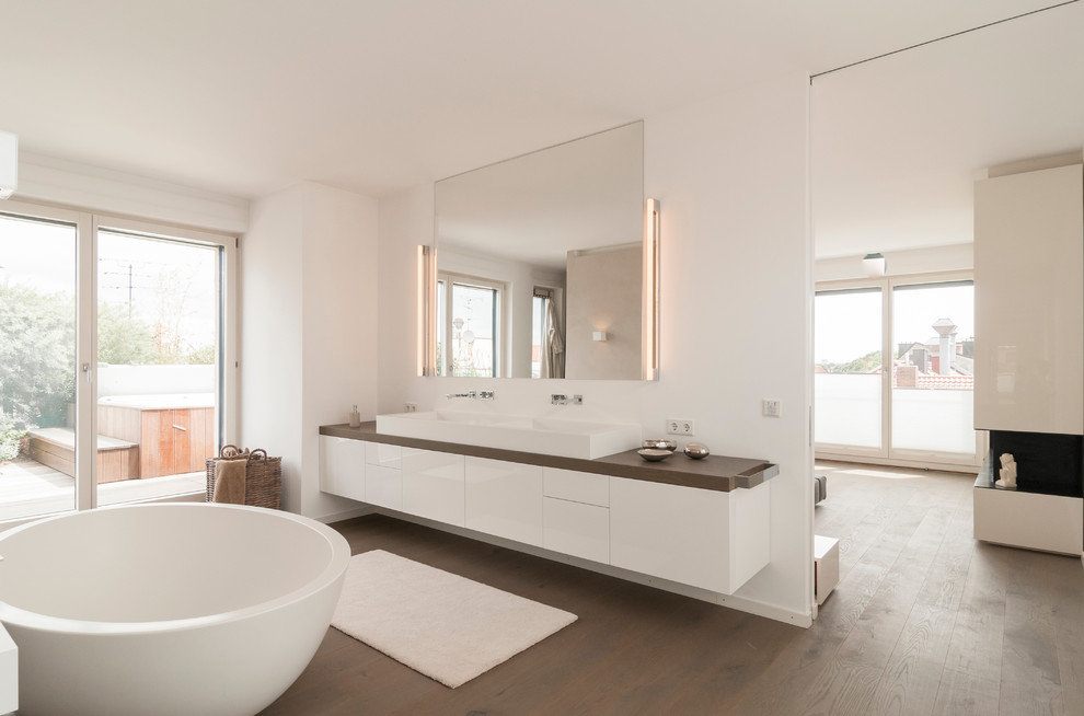 Пример оригинального дизайна: главная ванная комната среднего размера в современном стиле с плоскими фасадами, белыми фасадами, отдельно стоящей ванной, белыми стенами, паркетным полом среднего тона, настольной раковиной и столешницей из дерева