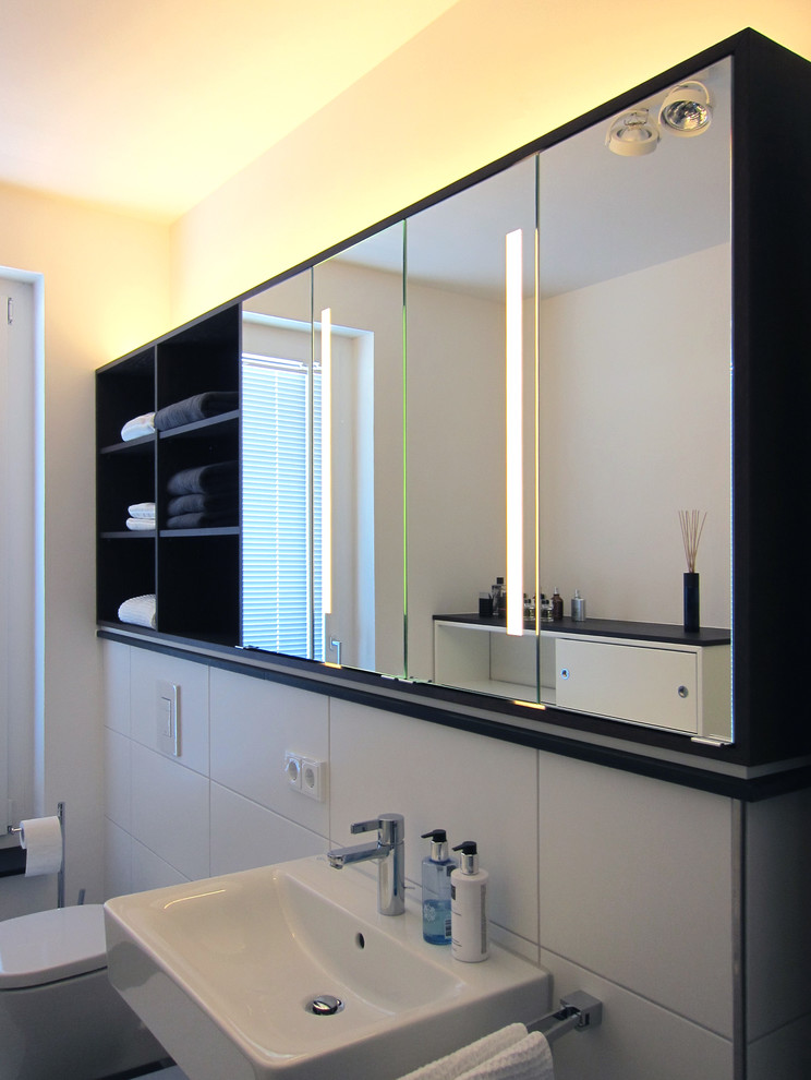 Photo of a medium sized contemporary bathroom in Cologne with flat-panel cabinets, white cabinets, a built-in bath, a shower/bath combination, a wall mounted toilet, white tiles, ceramic tiles, white walls, ceramic flooring, a wall-mounted sink, black floors and a hinged door.