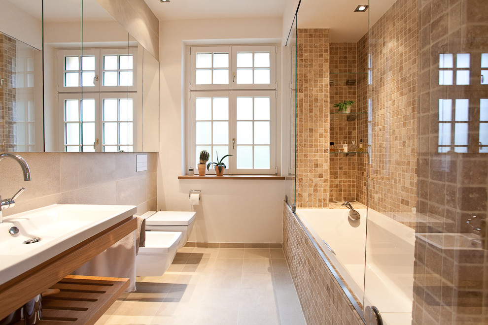 Foto på ett mellanstort vintage badrum, med ett badkar i en alkov, en dusch/badkar-kombination, en vägghängd toalettstol, beige kakel, brun kakel, mosaik, vita väggar, ett fristående handfat och träbänkskiva
