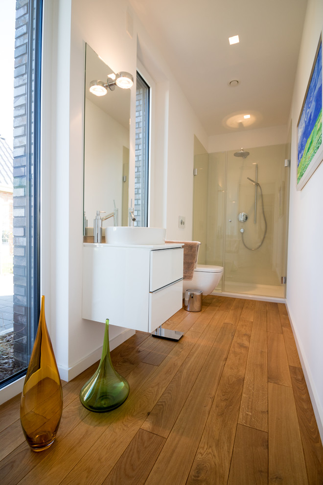 Cette image montre une petite douche en alcôve design avec un placard à porte plane, des portes de placard blanches, WC suspendus, un mur blanc, un sol en bois brun, une vasque et un sol marron.