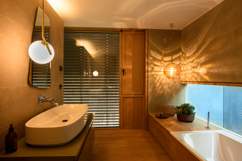 Cette image montre une salle de bain principale design de taille moyenne avec un placard à porte plane, des portes de placard grises, une baignoire posée, un carrelage beige, un mur gris, un sol en bois brun, une vasque, un sol marron et un plan de toilette gris.