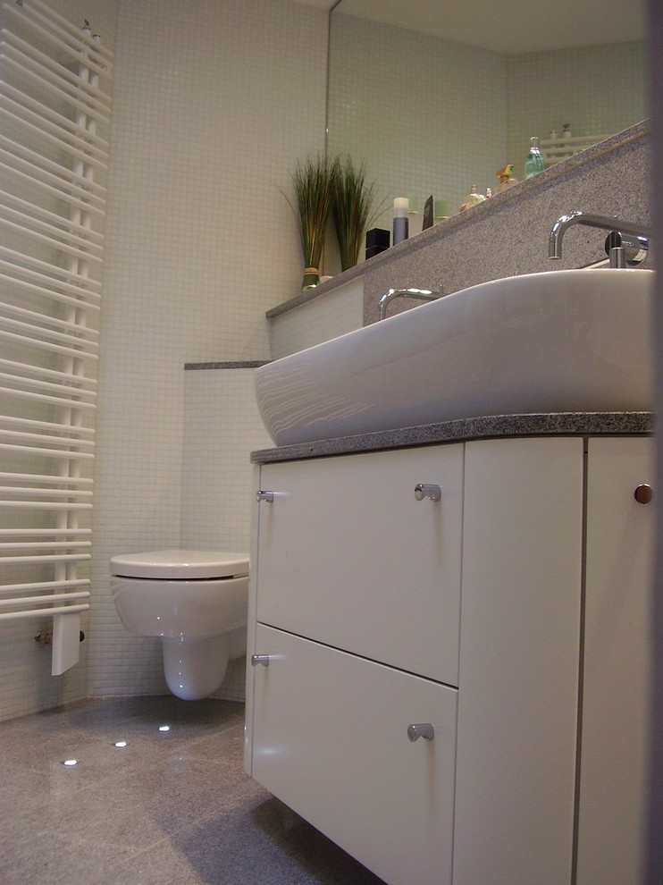 Cette photo montre une grande salle de bain principale tendance avec un placard à porte plane, des portes de placard blanches, une baignoire posée, un combiné douche/baignoire, WC suspendus, un carrelage blanc, un carrelage en pâte de verre, une vasque, un plan de toilette en granite, un sol gris, aucune cabine et un plan de toilette gris.