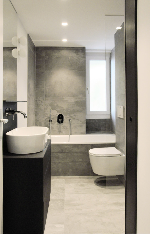 Cette image montre une petite salle de bain grise et blanche design avec un placard à porte plane, des portes de placard noires, une baignoire en alcôve, WC suspendus, un carrelage gris et une vasque.