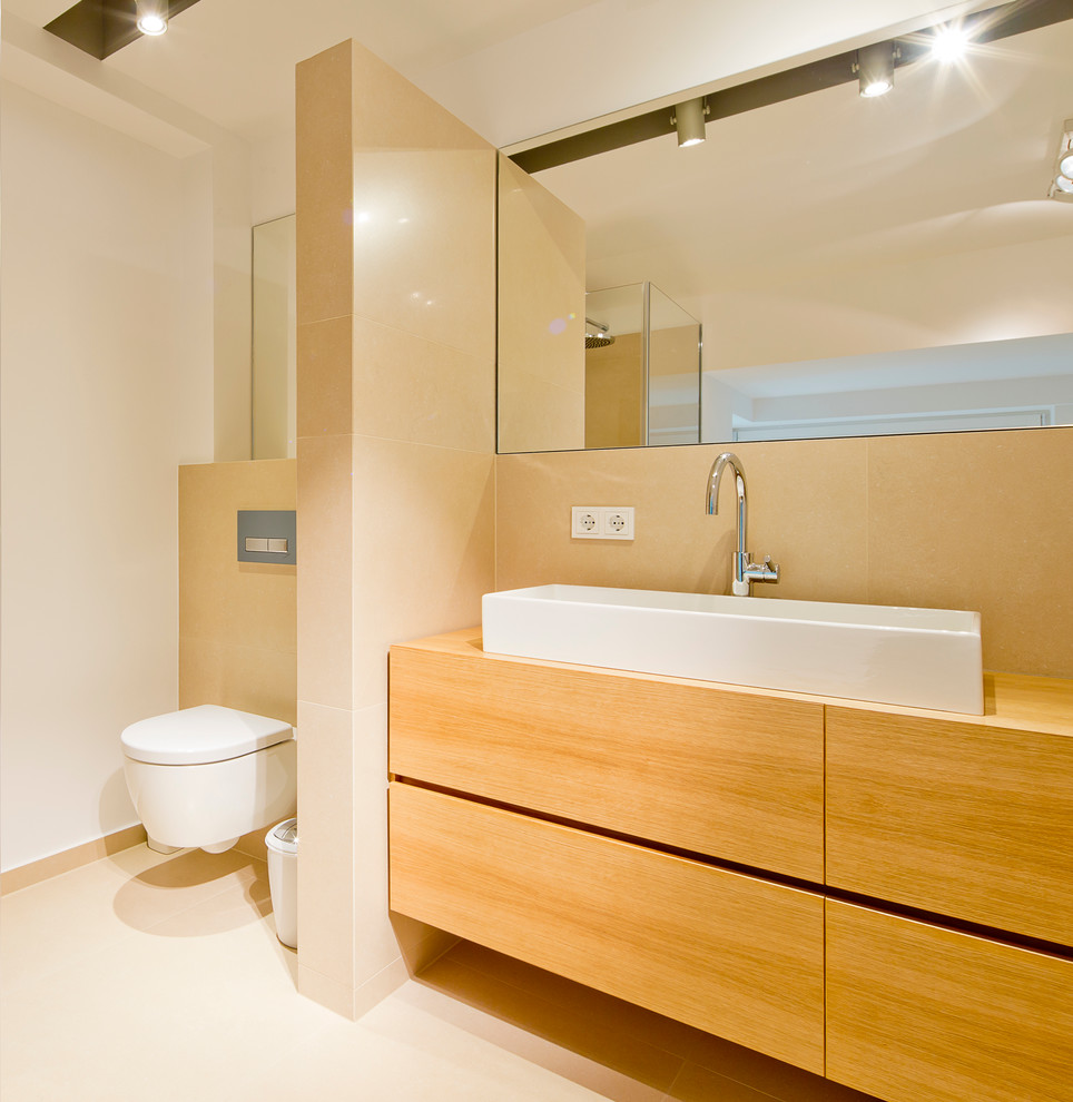 Cette image montre une grande salle de bain design en bois brun avec un placard à porte plane, WC suspendus, un carrelage beige, un mur blanc, une vasque, un plan de toilette en bois et un plan de toilette marron.