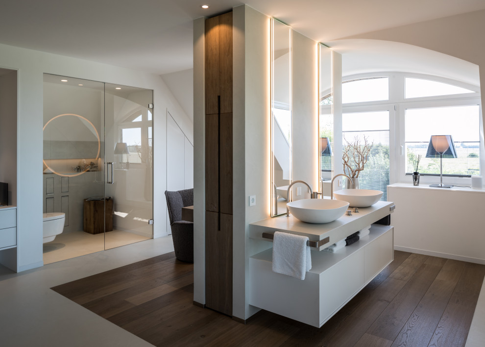 Photo of a contemporary bathroom in Bremen with flat-panel cabinets, white cabinets, white walls, dark hardwood flooring, a vessel sink, brown floors, white worktops, an enclosed toilet, double sinks and a floating vanity unit.