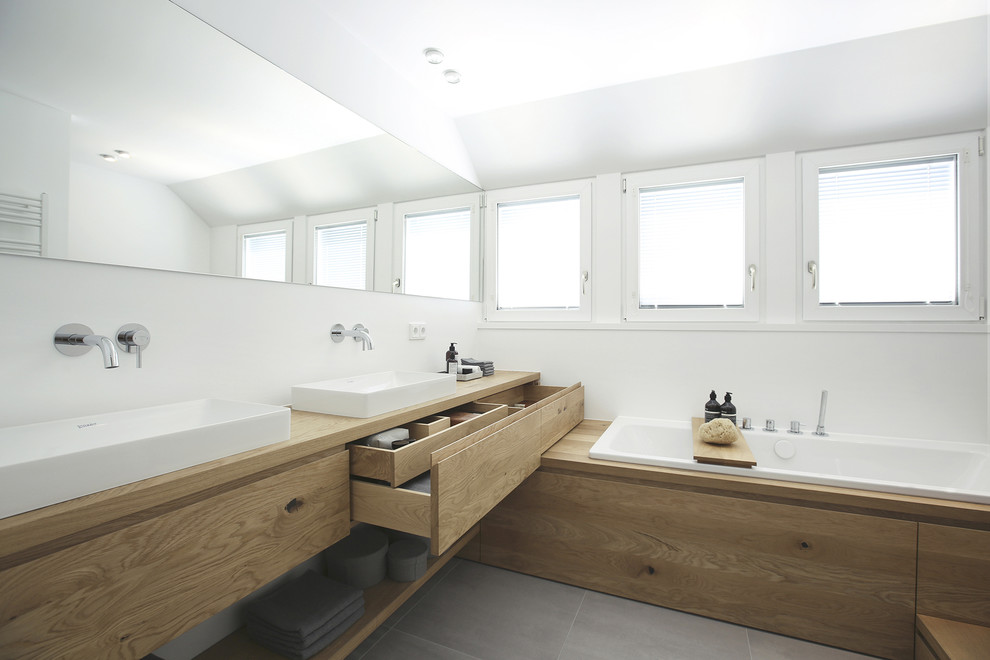 Medium sized contemporary bathroom in Frankfurt with flat-panel cabinets, light wood cabinets, a built-in bath, grey tiles, white walls, a vessel sink, wooden worktops and grey floors.