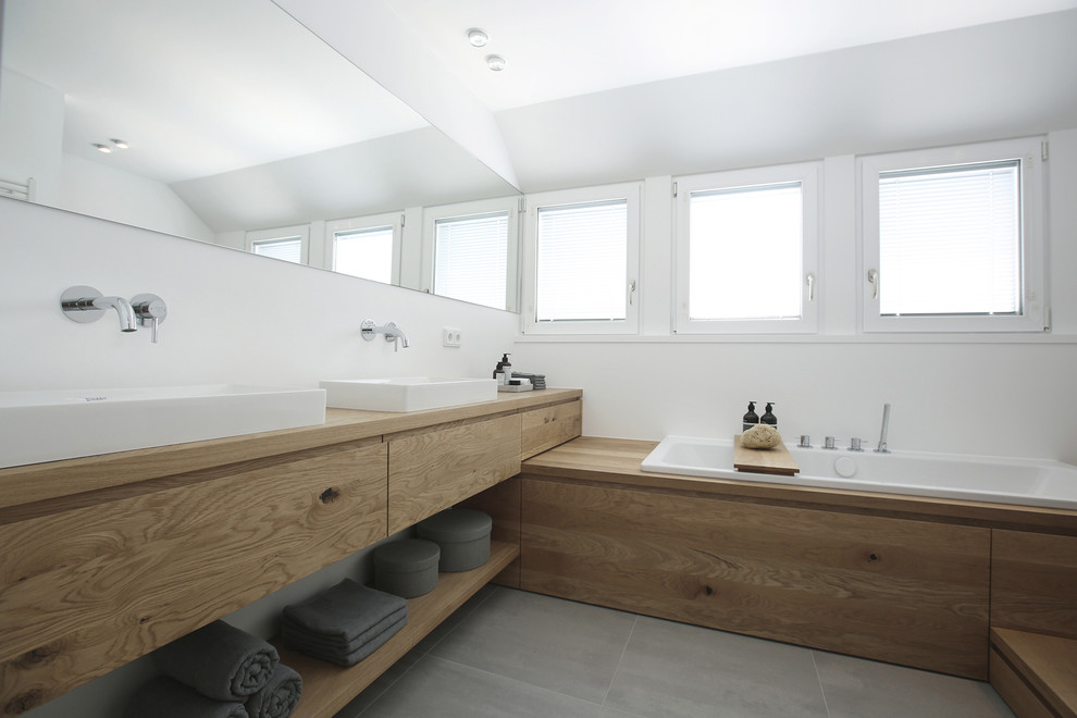Idéer för mellanstora funkis badrum, med släta luckor, skåp i ljust trä, ett platsbyggt badkar, grå kakel, vita väggar, ett fristående handfat, träbänkskiva och grått golv