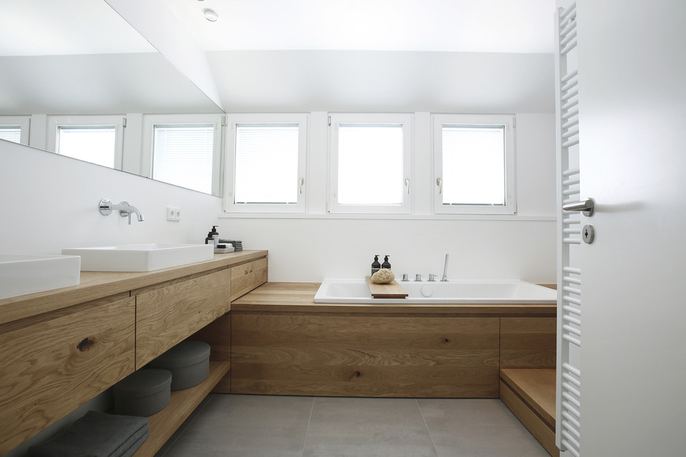 Modelo de cuarto de baño actual de tamaño medio con armarios con paneles lisos, puertas de armario de madera clara, bañera encastrada, baldosas y/o azulejos grises, paredes blancas, lavabo sobreencimera, encimera de madera y suelo gris