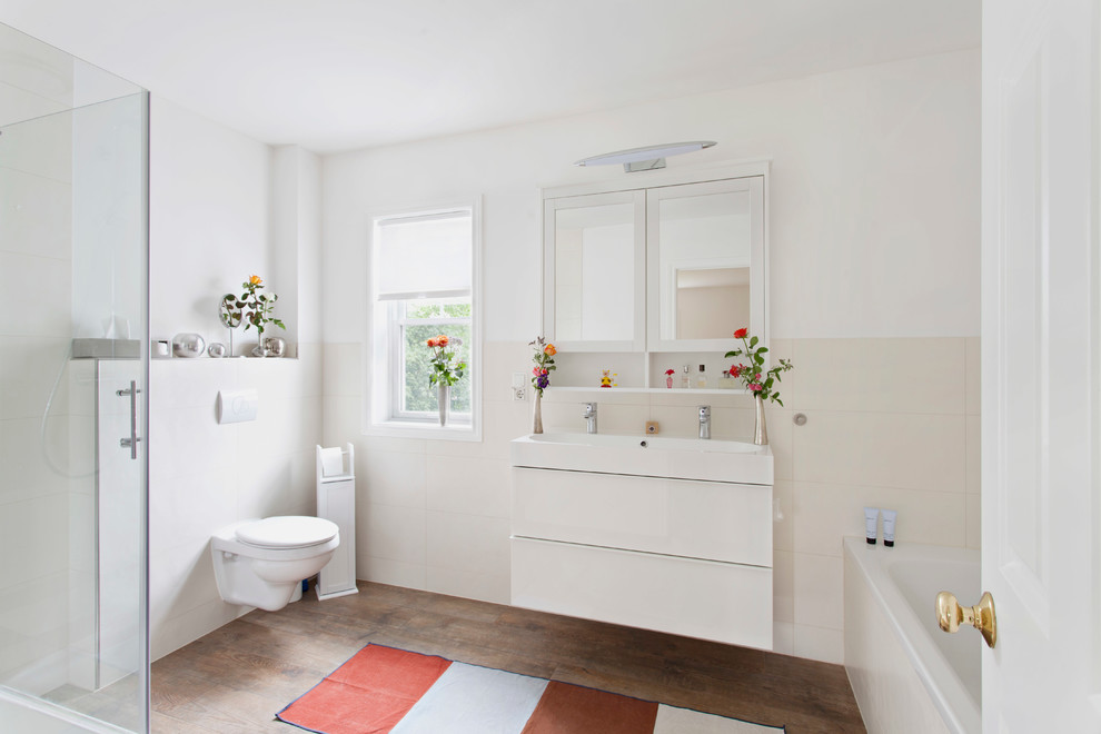 Idéer för ett mellanstort modernt badrum, med släta luckor, vita skåp, en vägghängd toalettstol, beige kakel, vita väggar, mellanmörkt trägolv, ett platsbyggt badkar, keramikplattor och ett avlångt handfat