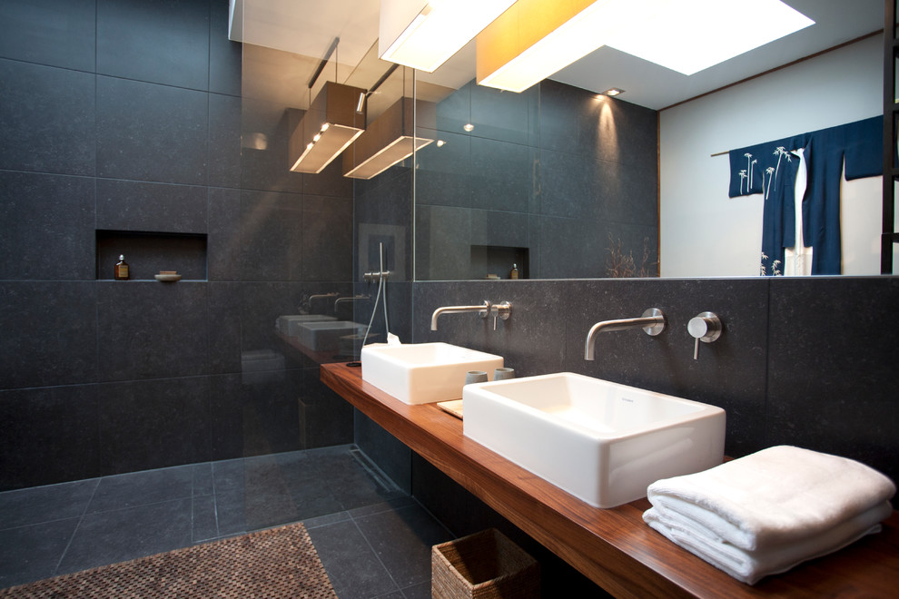 Bild på ett funkis badrum, med en vägghängd toalettstol, svart kakel, grå väggar, ett fristående handfat, träbänkskiva, ett fristående badkar och en öppen dusch
