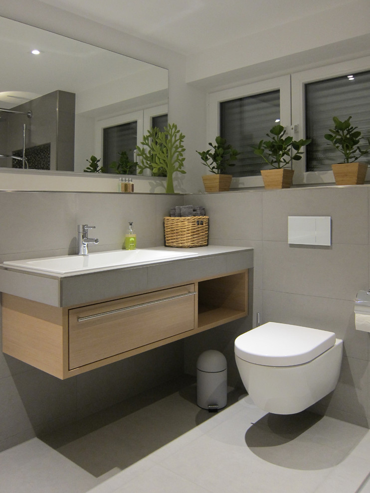 Foto på ett funkis badrum med dusch, med skåp i ljust trä, kaklad bänkskiva, grå kakel, keramikplattor, vita väggar, klinkergolv i keramik, ett nedsänkt handfat, en kantlös dusch och en vägghängd toalettstol