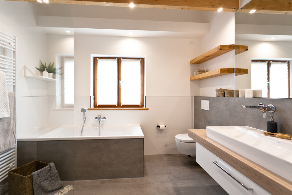 Modern inredning av ett mellanstort badrum med dusch, med släta luckor, vita skåp, ett platsbyggt badkar, en vägghängd toalettstol, brun kakel, cementkakel, vita väggar, cementgolv, ett fristående handfat och brunt golv