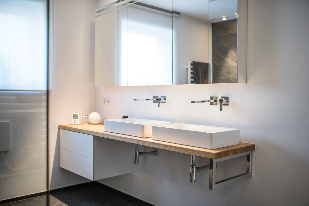 Cette image montre une salle de bain design de taille moyenne avec un placard à porte plane, des portes de placard blanches, un mur blanc, une vasque, un plan de toilette en bois, un sol noir et un plan de toilette marron.