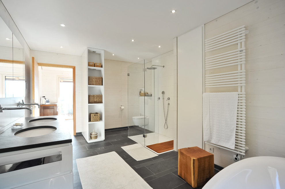 Expansive contemporary bathroom in Munich with white cabinets, a freestanding bath, a built-in shower, a wall mounted toilet, white tiles, ceramic tiles, white walls, slate flooring and a submerged sink.