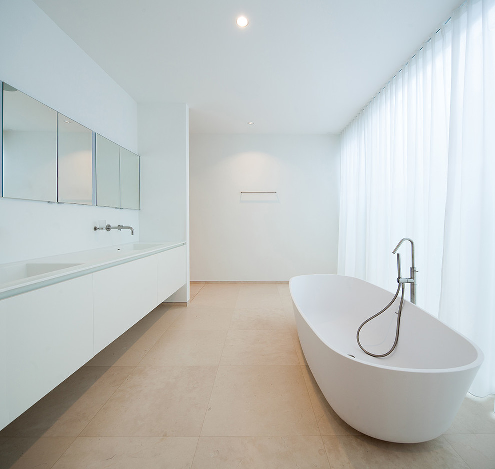 Diseño de cuarto de baño minimalista grande con bañera exenta, armarios con paneles lisos, puertas de armario blancas, paredes blancas, suelo de piedra caliza y lavabo integrado