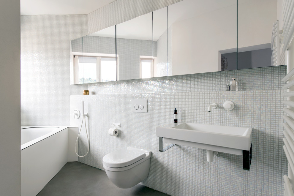 Inredning av ett modernt litet badrum, med ett undermonterat badkar, en vägghängd toalettstol, mosaik, vita väggar, ett väggmonterat handfat och betonggolv
