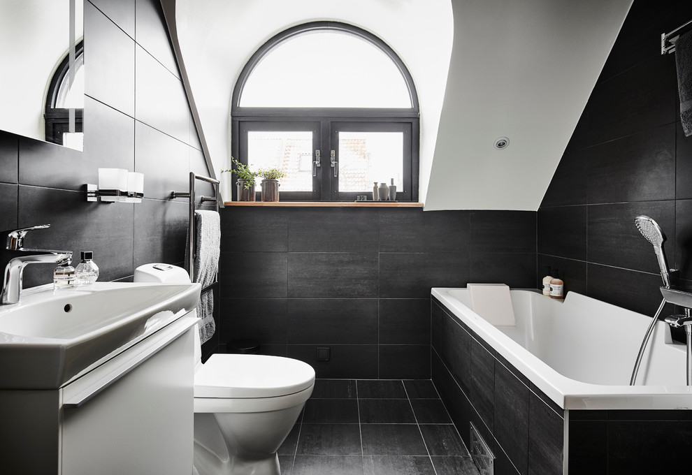 Photo of a large contemporary bathroom in Gothenburg with flat-panel cabinets, white cabinets, a submerged bath, a shower/bath combination, a one-piece toilet, ceramic tiles, black walls, limestone flooring, an integrated sink, black tiles and black floors.