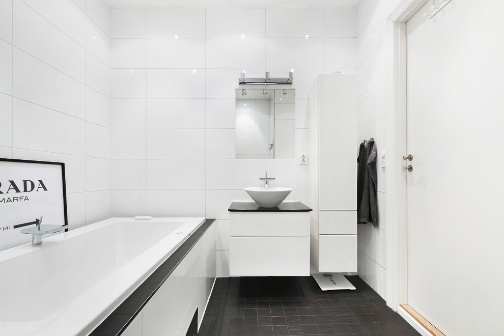 Réalisation d'une salle de bain nordique de taille moyenne avec un placard à porte plane, des portes de placard blanches, une baignoire posée, un carrelage blanc, un mur blanc, une vasque, un sol noir et un plan de toilette noir.