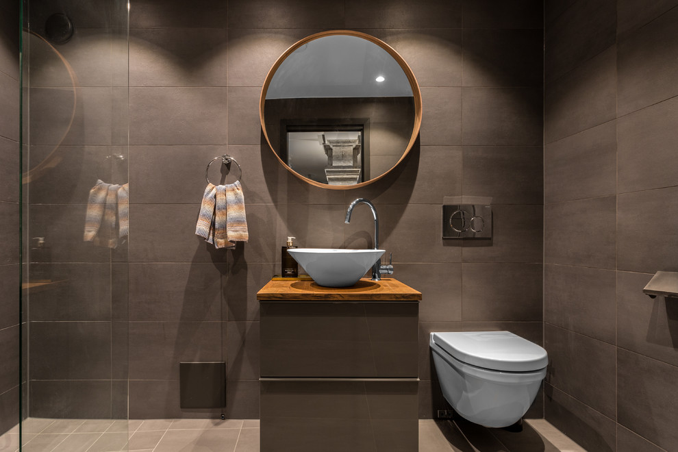 Idéer för att renovera ett mellanstort funkis brun brunt badrum med dusch, med släta luckor, bruna skåp, brun kakel, bruna väggar, ett fristående handfat, träbänkskiva, en kantlös dusch, en vägghängd toalettstol och grått golv
