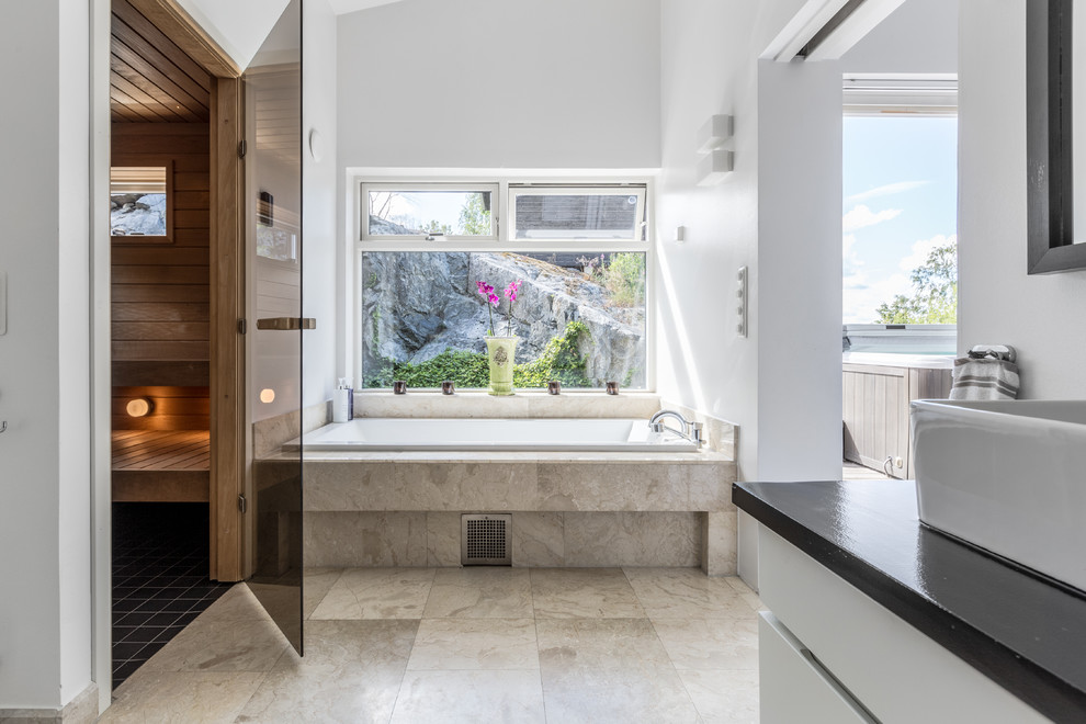Moderne Sauna mit flächenbündigen Schrankfronten, weißen Schränken, Einbaubadewanne, weißer Wandfarbe, Aufsatzwaschbecken, beigem Boden und schwarzer Waschtischplatte in Stockholm