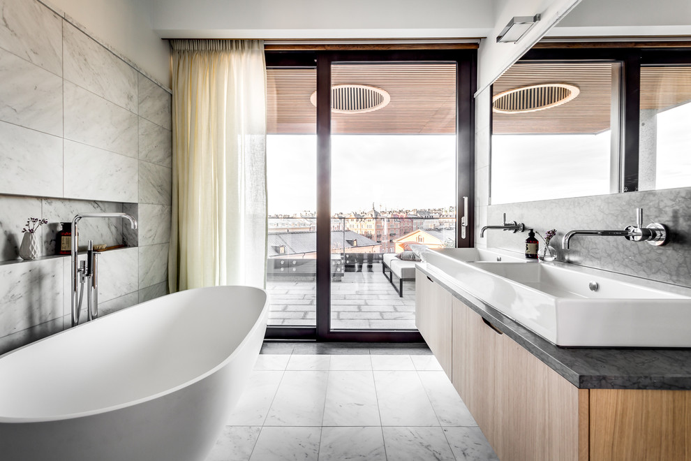 Inredning av ett modernt badrum, med släta luckor, skåp i ljust trä, ett fristående badkar, grå väggar, ett fristående handfat och grått golv