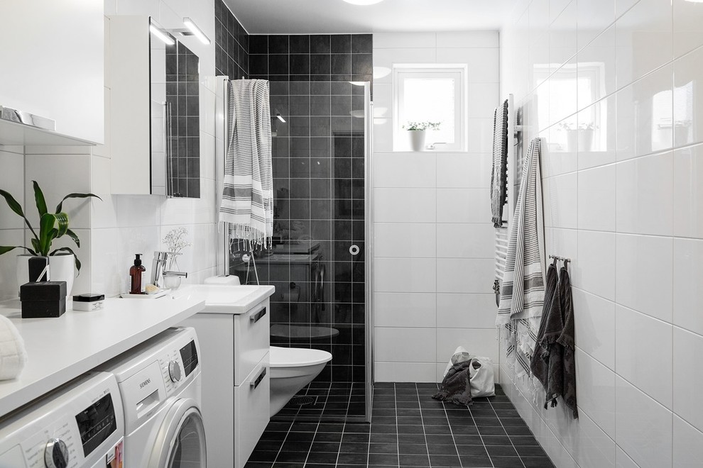 ヨーテボリにある北欧スタイルのおしゃれなバスルーム (浴槽なし) (フラットパネル扉のキャビネット、白いキャビネット、コーナー設置型シャワー、壁掛け式トイレ、モノトーンのタイル、コンソール型シンク、黒い床、開き戸のシャワー、洗濯室) の写真