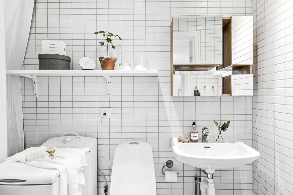 Foto på ett skandinaviskt badrum, med en toalettstol med hel cisternkåpa, vit kakel, vita väggar och ett väggmonterat handfat