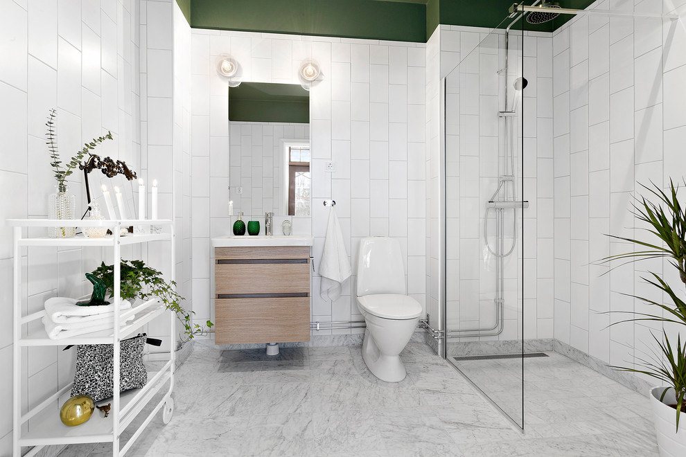 Skandinavisk inredning av ett badrum med dusch, med släta luckor, skåp i ljust trä, en hörndusch, en toalettstol med hel cisternkåpa, vit kakel, vita väggar, marmorgolv och flerfärgat golv