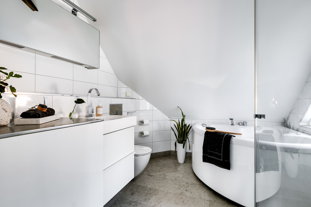 Modelo de cuarto de baño principal escandinavo con armarios con paneles lisos, puertas de armario blancas, jacuzzi, baldosas y/o azulejos blancos, paredes blancas, lavabo integrado, encimera de acero inoxidable, suelo gris y encimeras grises