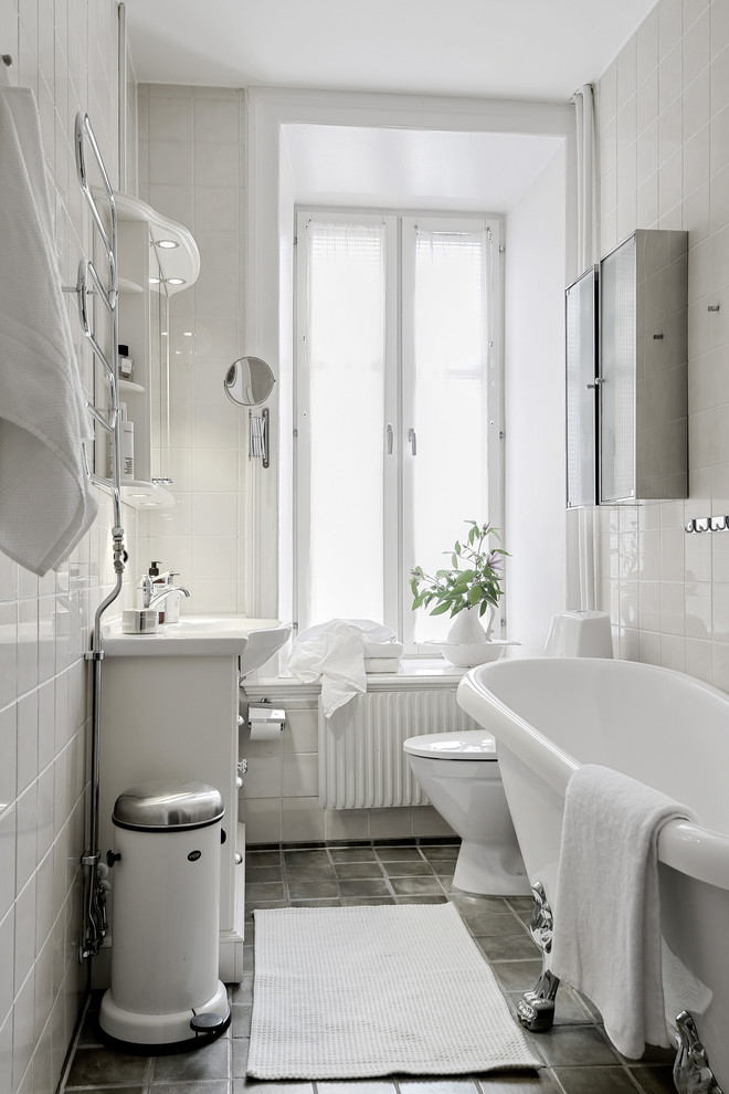 Inredning av ett nordiskt badrum, med vit kakel, vita skåp, ett badkar med tassar, vita väggar, ett konsol handfat och grått golv