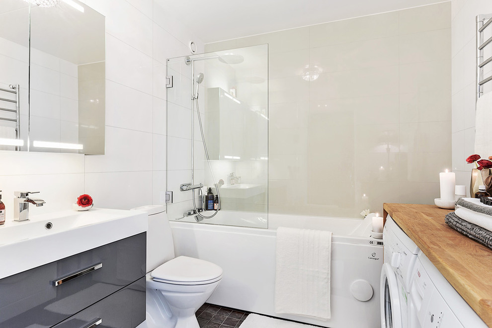 ストックホルムにあるお手頃価格の中くらいな北欧スタイルのおしゃれな浴室 (フラットパネル扉のキャビネット、グレーのキャビネット、ドロップイン型浴槽、シャワー付き浴槽	、分離型トイレ、ベージュのタイル、黒いタイル、白いタイル、白い壁、一体型シンク、磁器タイル、セラミックタイルの床、オープンシャワー、洗濯室) の写真
