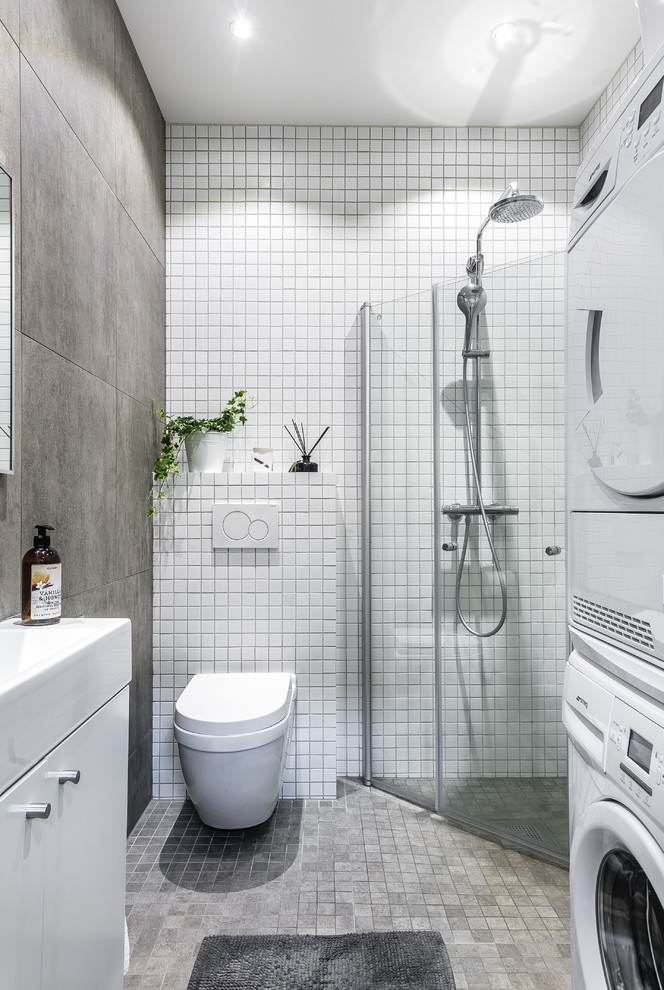 Foto på ett mellanstort nordiskt badrum med dusch, med en kantlös dusch, en vägghängd toalettstol, grå kakel, vit kakel, grått golv, släta luckor, vita skåp, mosaik, vita väggar, mosaikgolv och ett konsol handfat