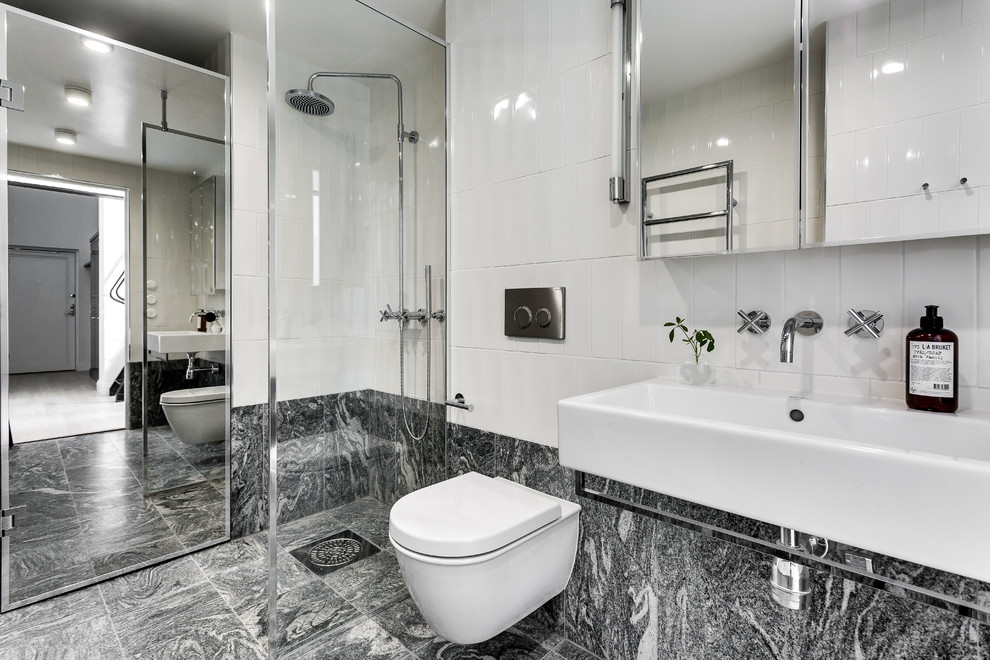 Bild på ett mellanstort nordiskt badrum med dusch, med en hörndusch, en vägghängd toalettstol, vit kakel, porslinskakel, ett avlångt handfat, vita väggar, marmorgolv och med dusch som är öppen