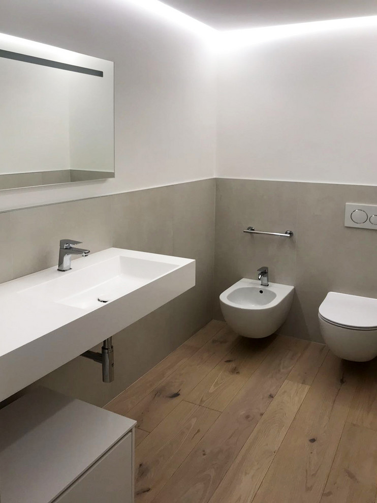 Exemple d'un WC et toilettes tendance de taille moyenne avec un placard à porte vitrée, des portes de placard grises, WC séparés, un carrelage gris, des carreaux de porcelaine, un mur blanc, un sol en bois brun, un lavabo suspendu, un plan de toilette en surface solide et un plan de toilette blanc.