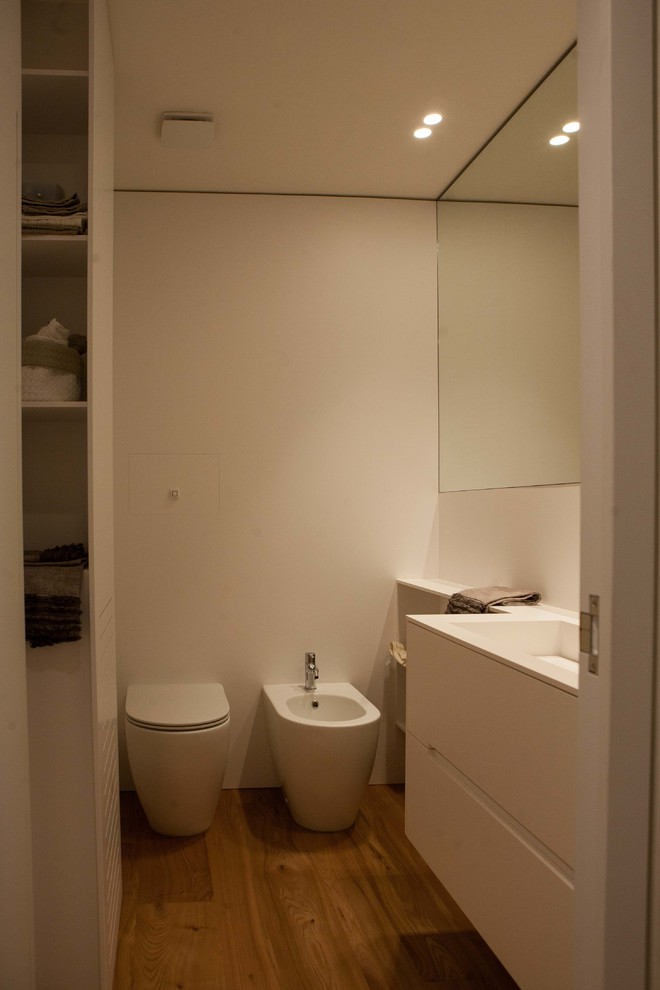Exemple d'un WC et toilettes tendance de taille moyenne avec des portes de placard blanches, un carrelage blanc, un mur blanc, parquet clair, un lavabo intégré, un plan de toilette en surface solide, un sol marron et un plan de toilette blanc.