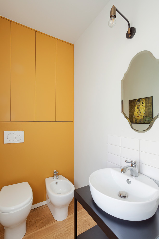 Cette photo montre un WC et toilettes éclectique avec un placard sans porte, des portes de placard noires, un carrelage blanc, un mur jaune, une vasque, un plan de toilette noir, WC à poser, un sol en bois brun et un sol beige.