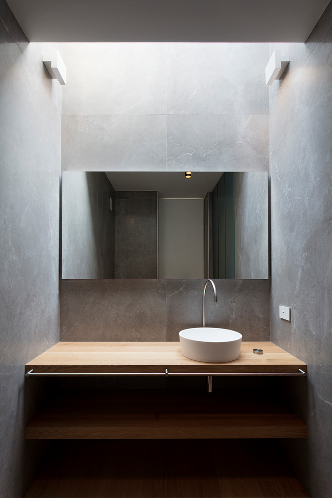 Cette image montre un WC et toilettes minimaliste en bois clair de taille moyenne avec un placard sans porte, un carrelage gris, un mur gris, une vasque et un plan de toilette en bois.