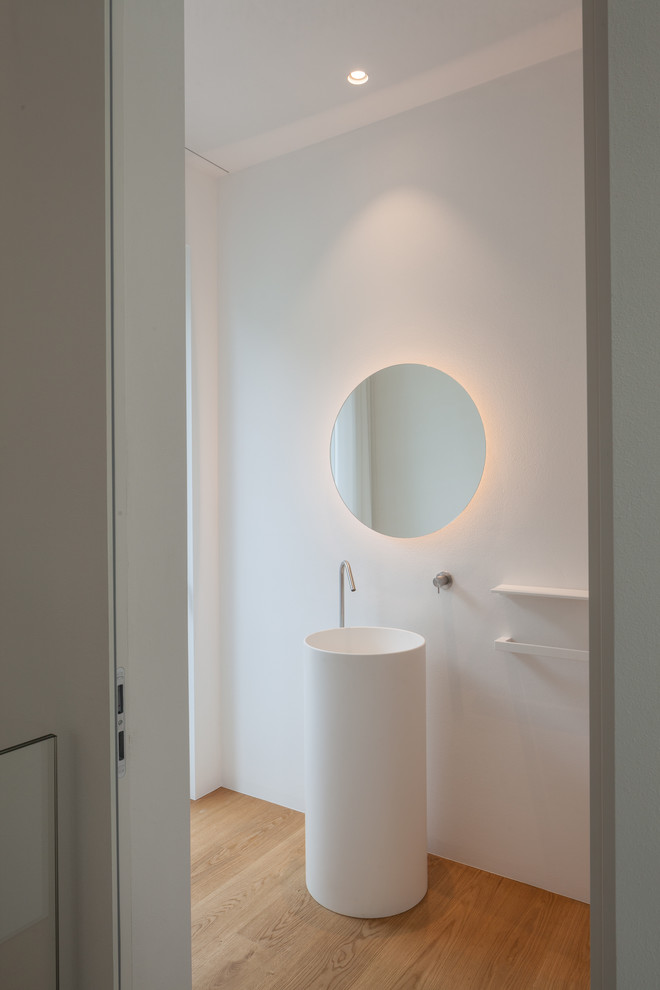 Foto på ett funkis vit toalett, med en vägghängd toalettstol, ljust trägolv, ett piedestal handfat och bänkskiva i akrylsten