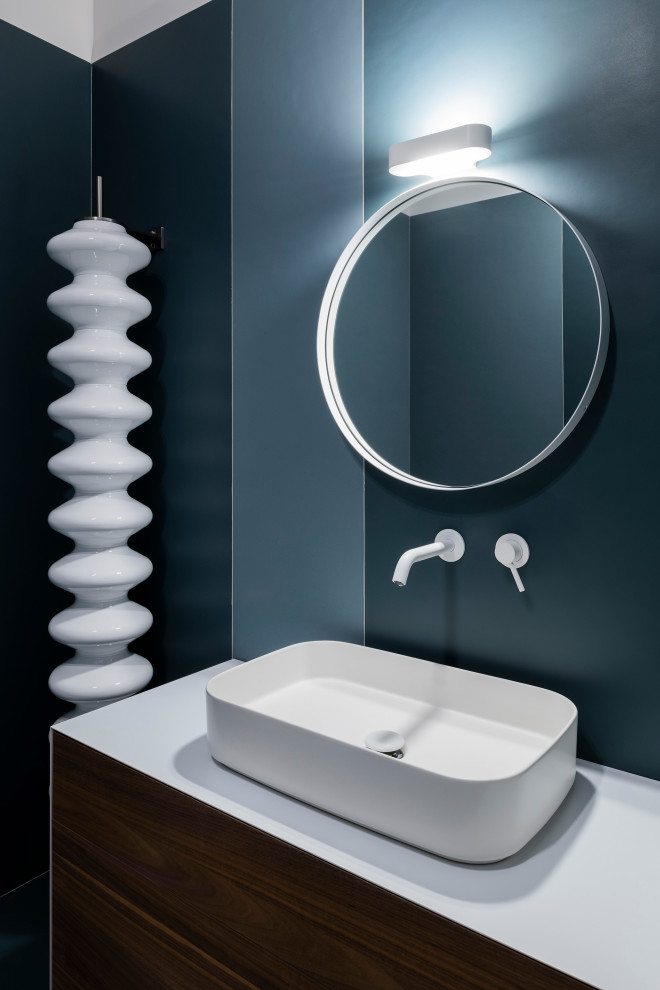 Cette image montre un WC et toilettes design en bois brun de taille moyenne avec un placard à porte plane, un mur bleu, une vasque, un plan de toilette blanc et meuble-lavabo encastré.
