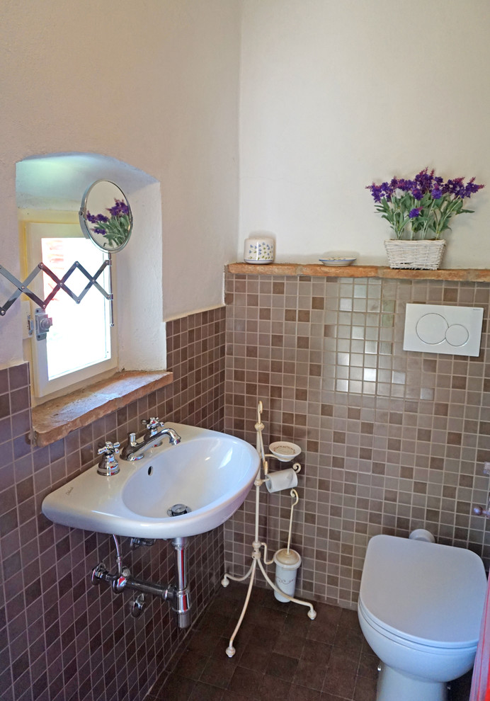 Пример оригинального дизайна: маленький туалет в стиле кантри с унитазом-моноблоком, коричневой плиткой, керамической плиткой, белыми стенами, полом из керамической плитки, подвесной раковиной и коричневым полом для на участке и в саду