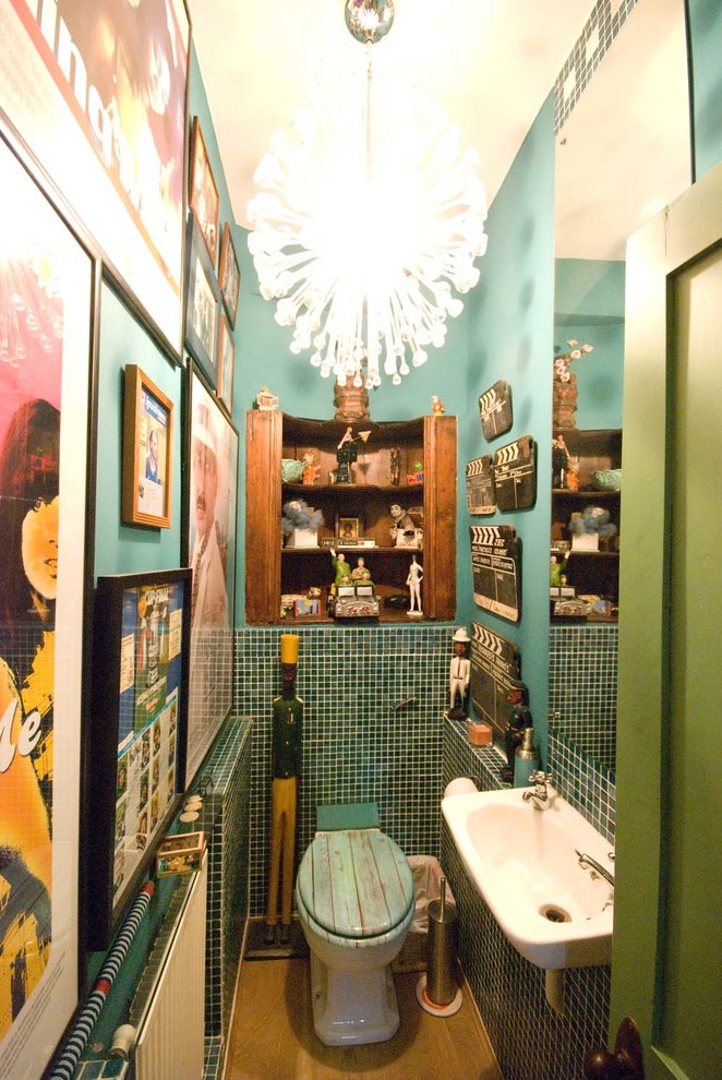 Inredning av ett eklektiskt litet toalett, med öppna hyllor, skåp i ljust trä, en toalettstol med hel cisternkåpa, grön kakel, mosaik, gröna väggar, ljust trägolv och ett väggmonterat handfat