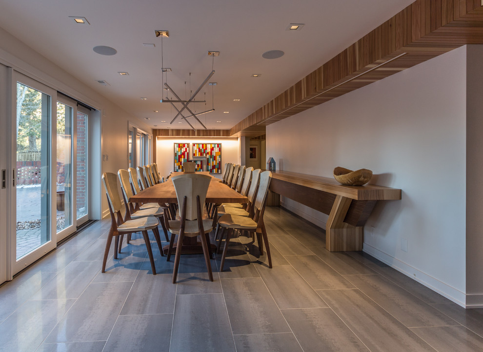 This is an example of a contemporary dining room with white walls, porcelain flooring, a standard fireplace, a tiled fireplace surround and grey floors.