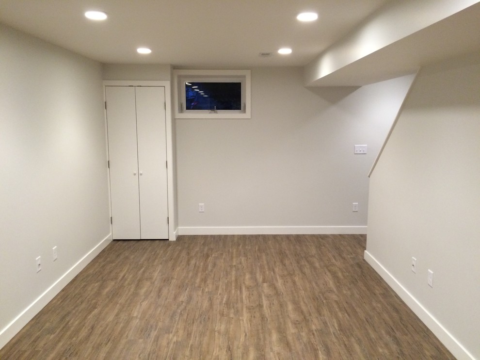 Cette image montre un sous-sol minimaliste semi-enterré et de taille moyenne avec un mur blanc, un sol en bois brun, aucune cheminée et un sol marron.
