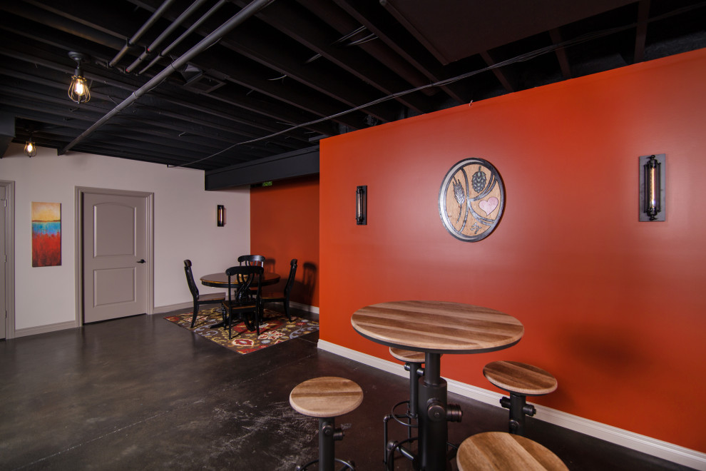 Cette photo montre un grand sous-sol industriel avec un bar de salon, un mur orange, sol en béton ciré et poutres apparentes.